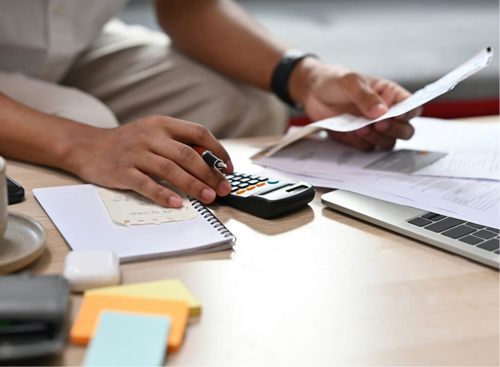 Close up man sitting on a couch in a living room looking at papers and using a calculator and a laptop.